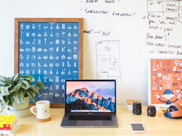 Laptop computer on desk surrounded by design materials, calendars, and creative workspace items.