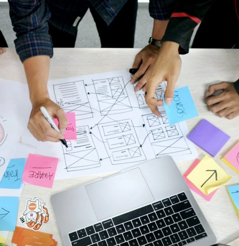 Team collaborating over wireframe sketches and sticky notes on white table with laptop nearby.
