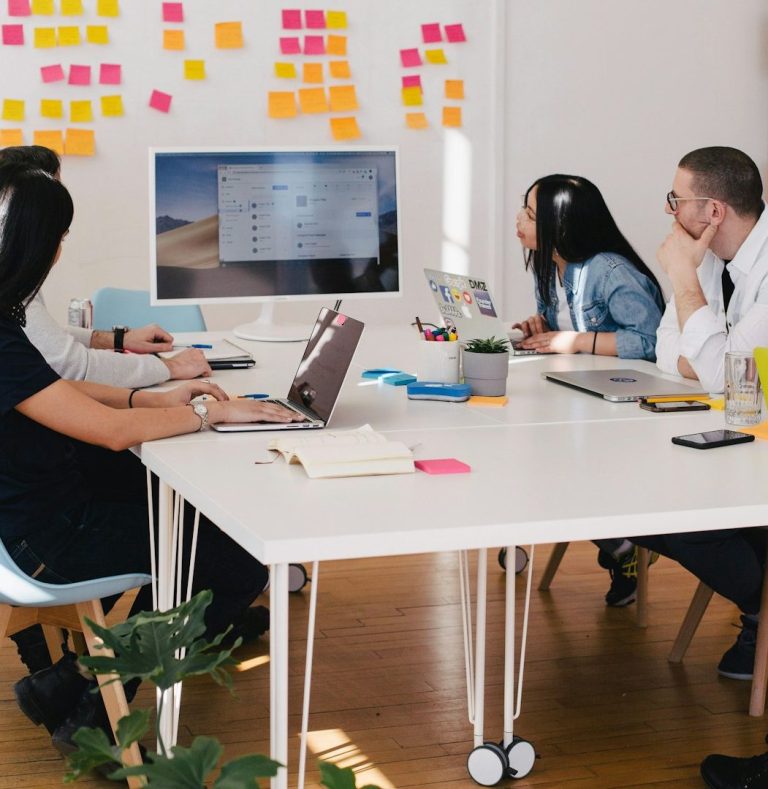 Team meeting around conference table with laptops, colourful sticky notes on wall, and monitor display.