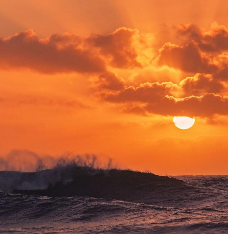 Dramatic orange sunset sky with sun visible through clouds over silhouetted breaking waves.
