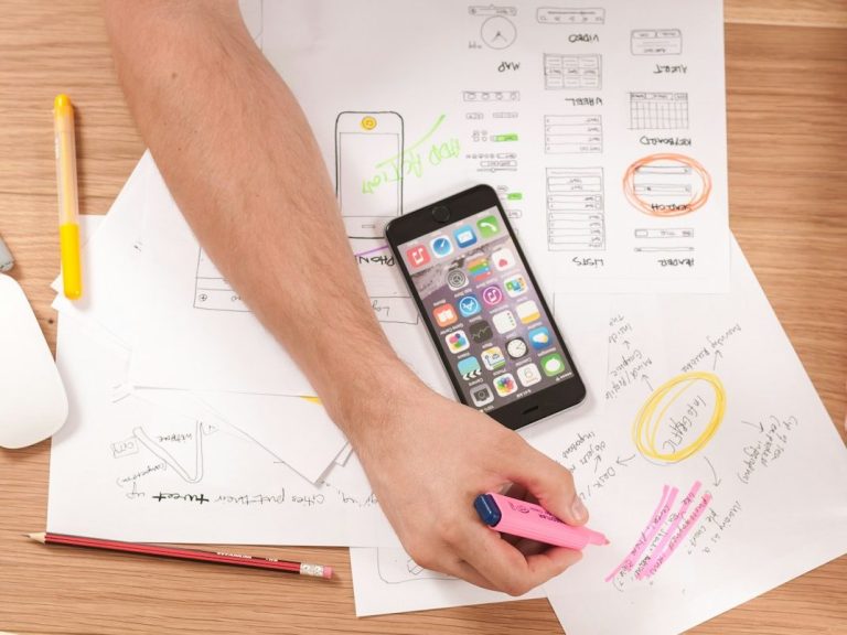 Hand reaching for smartphone on desk covered with papers, charts, and planning documents.