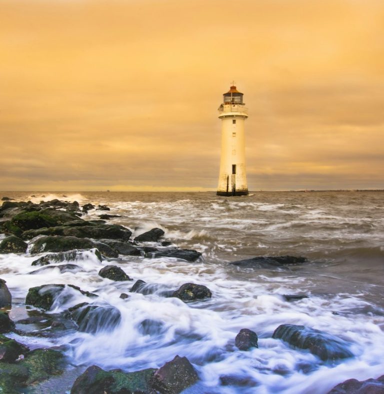 White lighthouse on rocky coastline with waves crashing over dark rocks at golden hour.